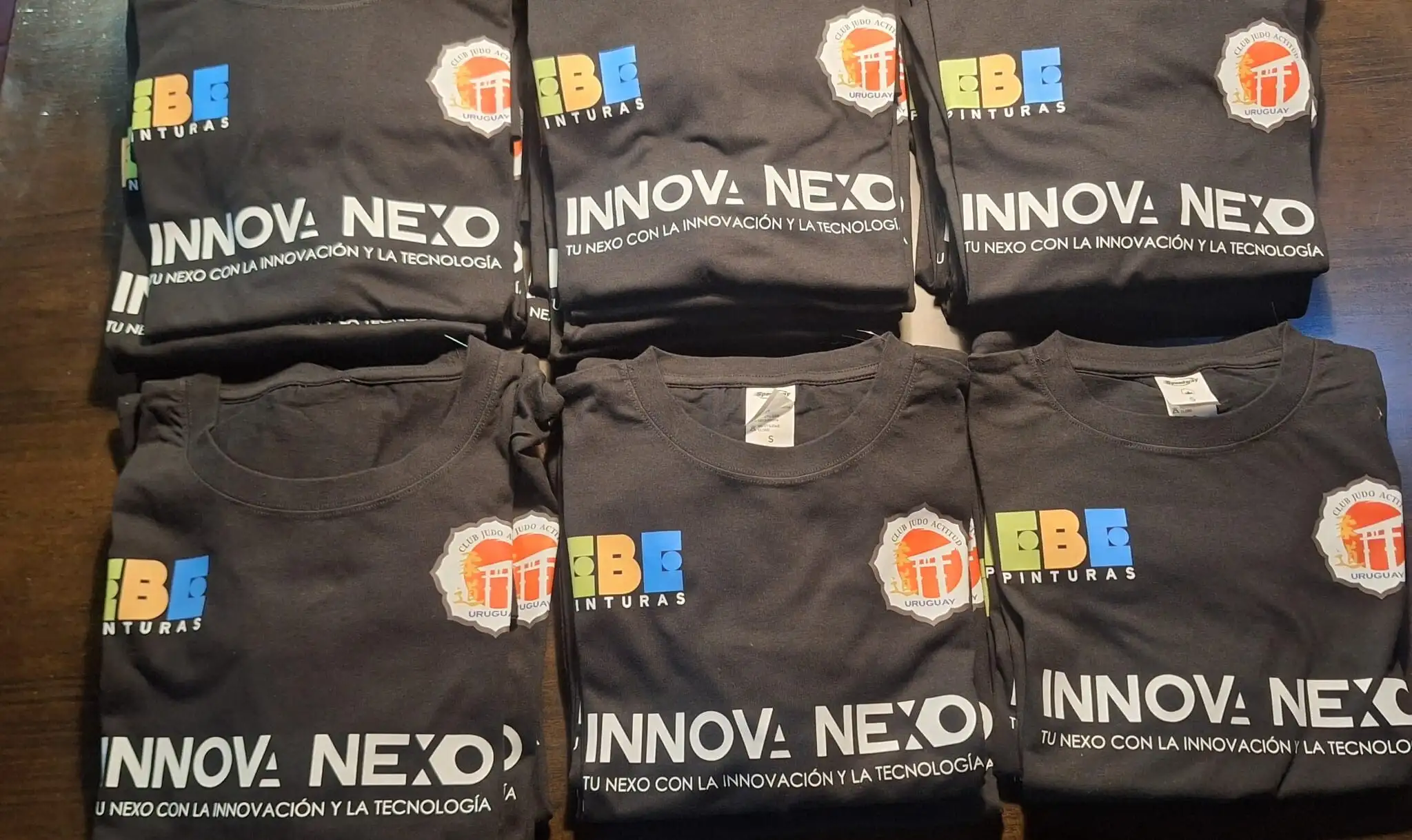 Pila de remeras negras dobladas con los logos estampados a color de Innova Nexo, EBE Pinturas y Club Judo Actitud