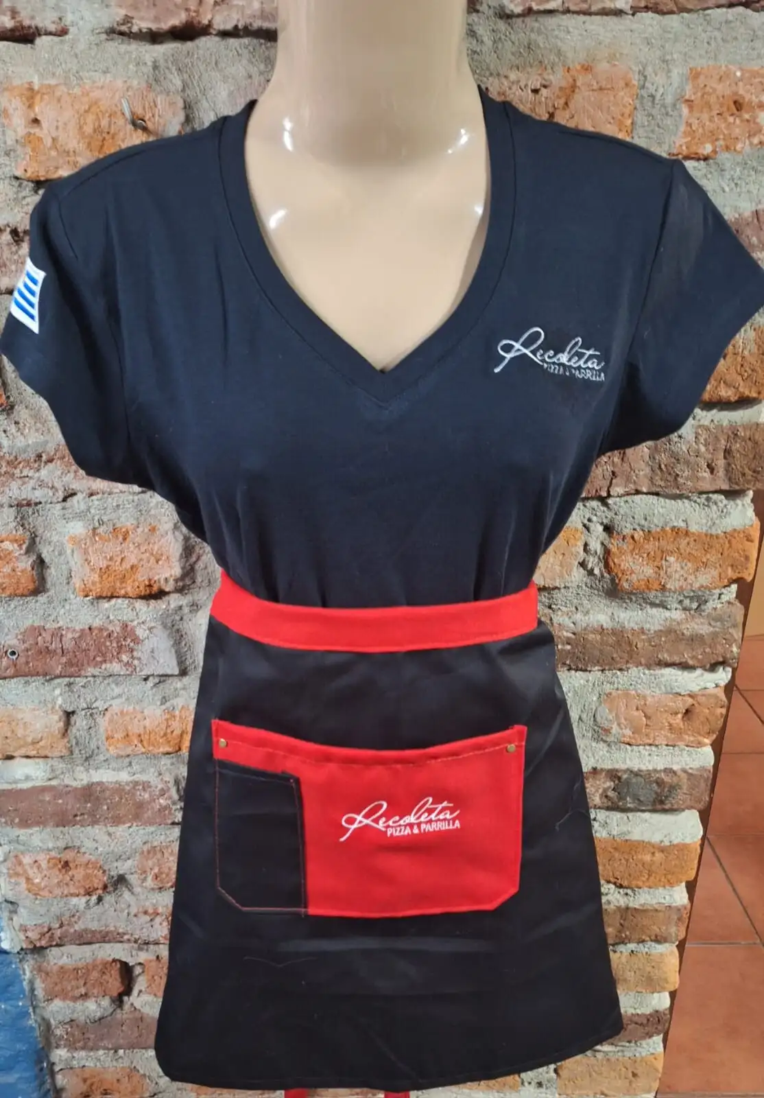 Remera azul con la bandera uruguaya en la manga y delantal negro con bolsillo rojo, ambos con el logo de Recoleta Pizza & Parrilla bordado